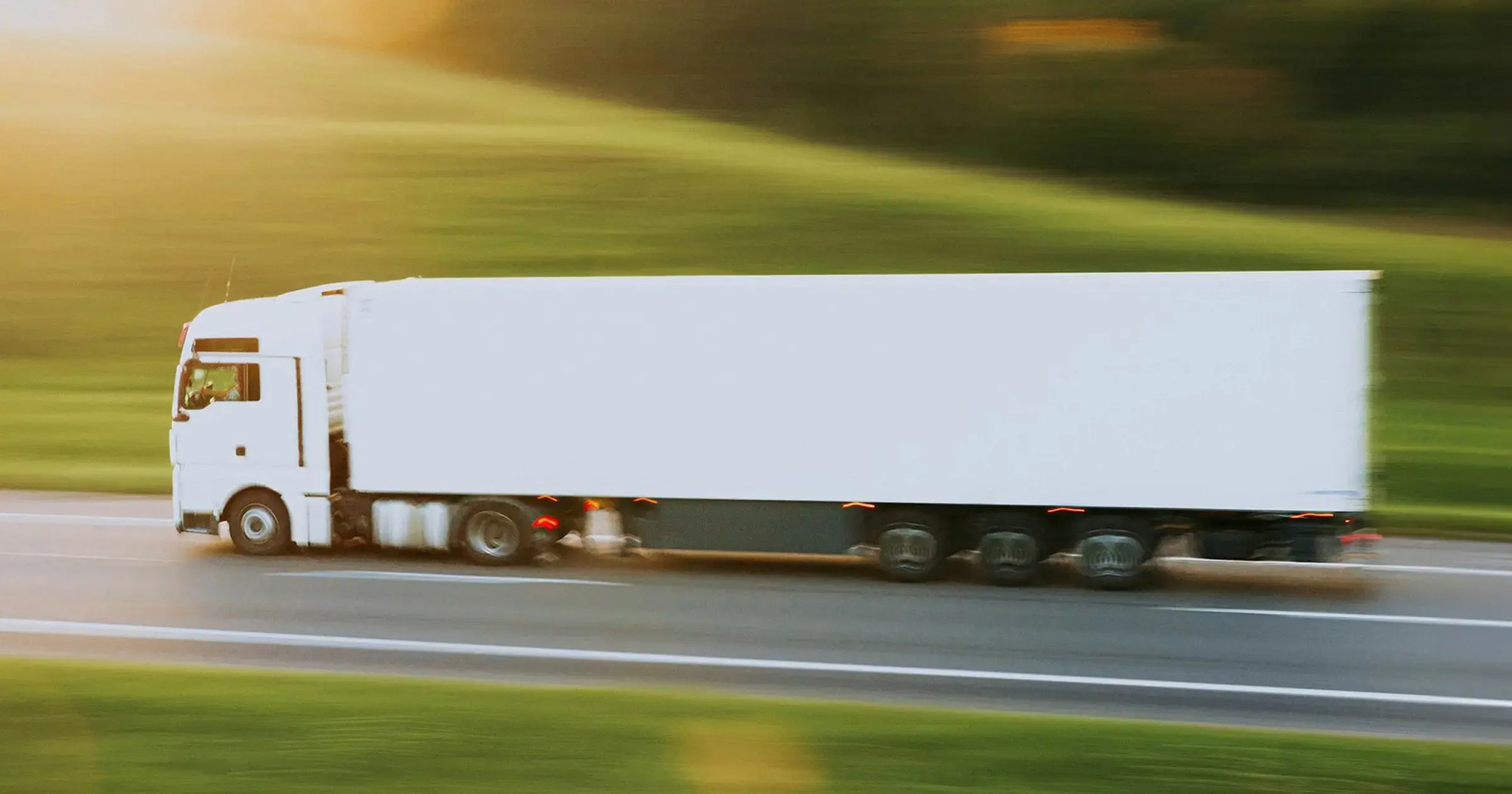 White transport truck driving at high speed on a highway with blurred green landscape in the background at sunset