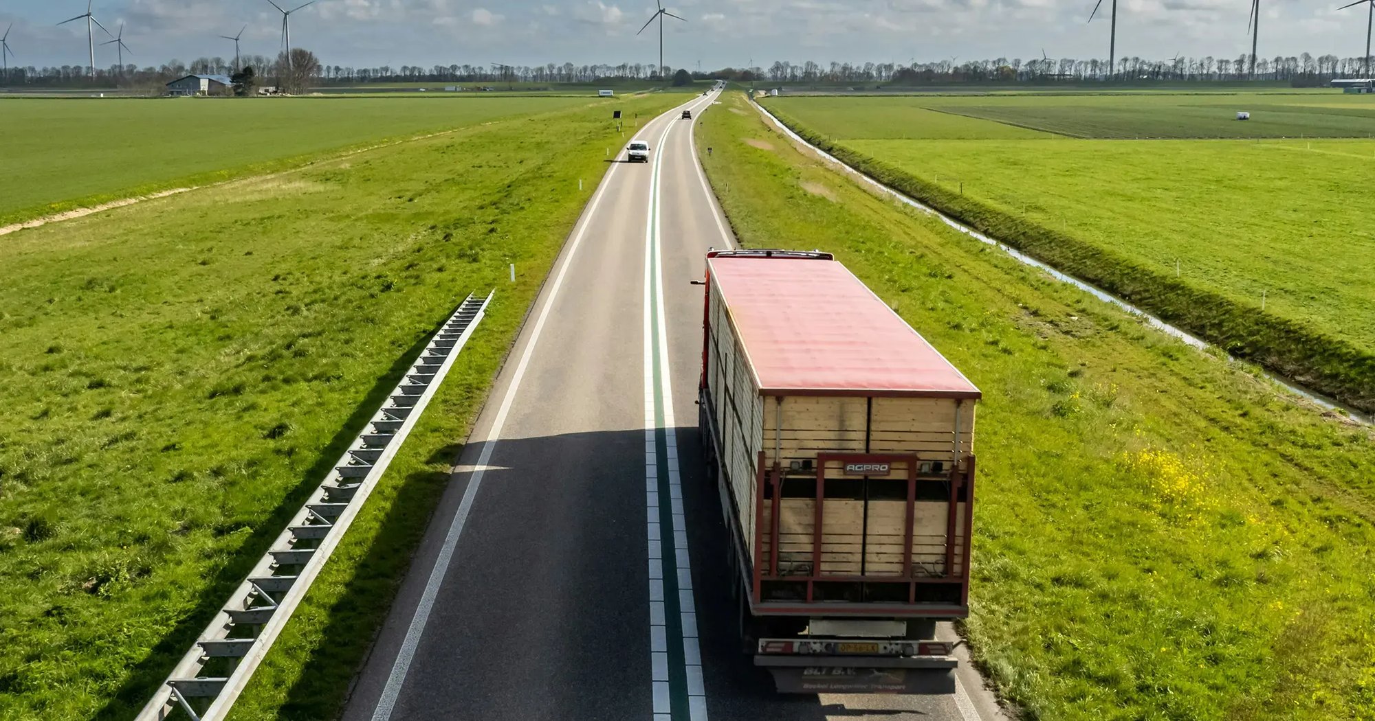 Transport truck driving on a rural road through green fields with wind turbines in the background